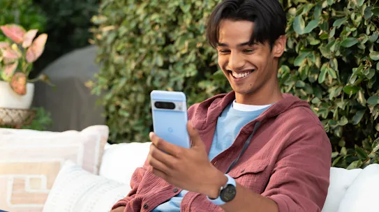 A man sitting on a patio outdoors while looking at a smartphone and smiling.