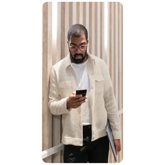 A man with a beard and glasses stands against a wall with vertical wooden slats. He is wearing a white t-shirt, a cream-colored textured button-down shirt, and black pants. He is looking down at a smartphone in his hands and holding a laptop under his arm.