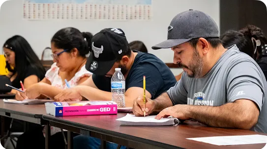 A man wearing a gray cap and t-shirt focused on writing in a notebook during a class, with a pink GED test preparation book sitting on the desk alongside other studying students.