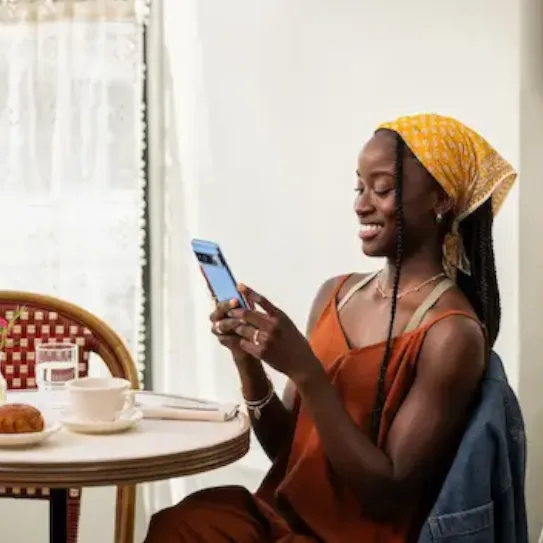 A person with long braids, wearing a yellow headscarf and a rust-coloured dress, sits at a table and smiles while using a smartphone. The setting appears to be a bright cafe or restaurant.