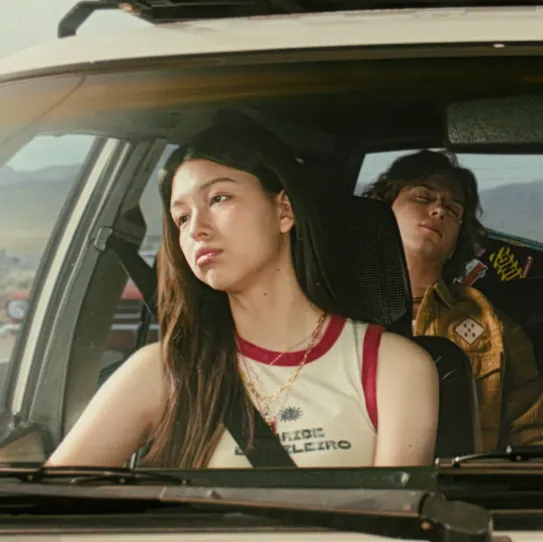 A group of friends are on a road trip through the desert. One of them uses their Samsung Galaxy smartphone to find a song by typing "Find the song that goes boom da la and play it." Gemini on the phone identifies the song as "BOOMPALA" by LE SSERAFIM and starts playing it through Spotify.