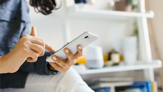 A person's hand holding a white smartphone and tapping the screen while sitting in a brightly lit room.