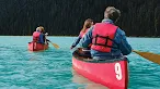 People wearing red life jackets paddle red canoes across a vibrant turquoise lake bordered by a dense evergreen forest.