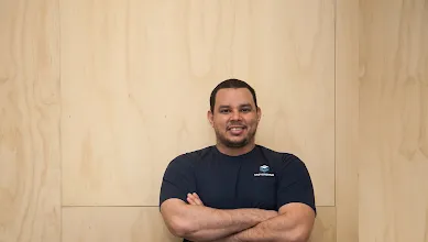 Homem em pé, de braços cruzados, em frente a fundo de madeira.