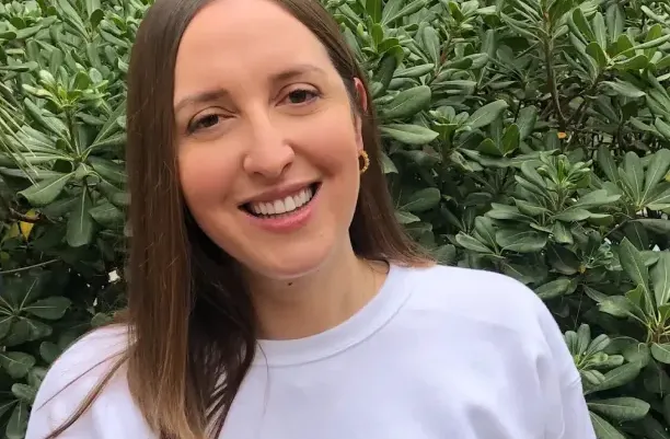Mujer sonriente de pie al aire libre, frente a un follaje verde, vistiendo una sudadera de color claro con el logotipo de Bounsel.