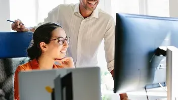 A woman in an orange top and a man in a white button-down shirt, both wearing glasses, smile while looking at a large computer monitor in a bright, sunlit office. This photo represents the 'Small business bAIsics' AI training program by the U.S. Chamber of Commerce, designed to help small business owners adopt AI tools.
