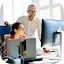 A woman in an orange top and a man in a white button-down shirt, both wearing glasses, smile while looking at a large computer monitor in a bright, sunlit office. This photo represents the 'Small business bAIsics' AI training program by the U.S. Chamber of Commerce, designed to help small business owners adopt AI tools.