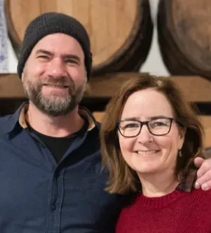 A smiling man in a dark beanie and blue button-down shirt stands next to a smiling woman with glasses and a red sweater, with wooden barrels in the background.
