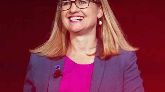 A medium portrait of Kate Gallego, the Mayor of Phoenix, Arizona. She is smiling and wearing glasses, a pink top, and a blue blazer with a small lapel pin. The background is a solid, warm red, suggesting she is speaking at an event or on a stage.
