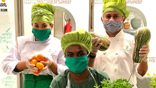 Three chefs in masks and white robes are holding up fresh produce for the camera.