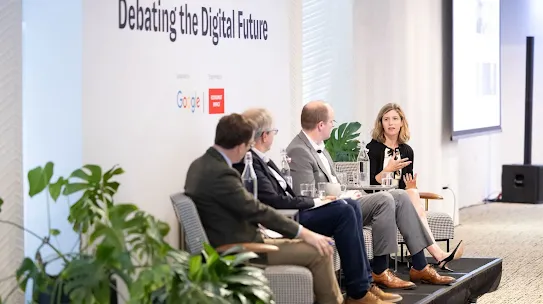 A panel of four experts sitting on a stage in front of a backdrop that reads "Debating the Digital Future." The stage features the Google and Economist Impact logos. A female panelist on the right is speaking and gesturing, while three male panelists listen. Large Monstera plants decorate the front of the stage.