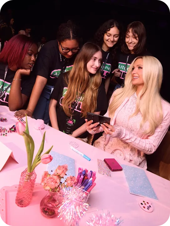 Paris Hilton in a pink dress sitting at a table, smiling at a group of girls in black "Android" shirts who are showing her something on a folding smartphone.