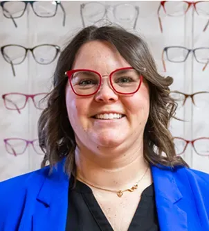 A portrait of an optometrist wearing red-rimmed glasses and a bright blue blazer, smiling in front of a display of various eyeglass frames in an Oklahoma vision care office.