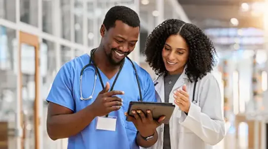 Two smiling healthcare professionals, a man in blue scrubs and a woman in a white lab coat, both with stethoscopes around their necks, looking at a digital tablet together in a bright hospital hallway.
