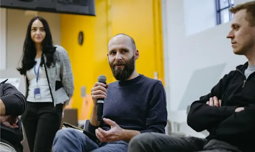 A man with a beard is speaking into a microphone. Two men and a woman are looking at him, smiling.