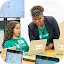 An instructor wearing a blazer over a green Google 4-H t-shirt assists a young girl at a laptop during a technology workshop.