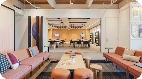 A modern corporate office lounge space featuring mid-century style orange couches, textured throw pillows, low coffee tables, and round stools. In the background, a large open partition leads into a brightly lit workshop or conference room filled with collaborative tables and chairs.