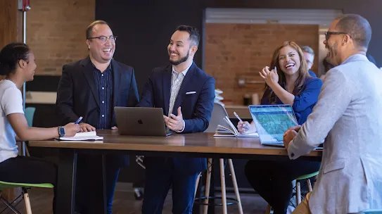 Innovare co-founders AJ DeLeón, Fernando DeLeón, and Nicholas Freeman laughing and collaborating with team members around a high-top office table with laptops.