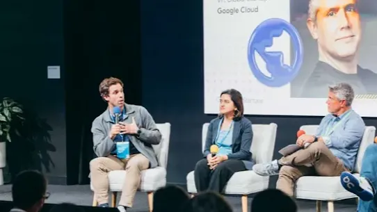 Three panelists are seated on stage in white chairs during a Google for Startups Gemini Startup Forum. Behind them, a large screen displays "Moderated by Darren Mowry, VP, Global Startups Google Cloud." One panelist is speaking into a microphone while an audience watches from the foreground, illustrating a live collaborative tech event.