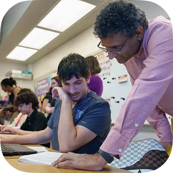 A male professor with glasses and a pink shirt leans over a desk to guide a male student working on a laptop. They are reviewing a document together in a brightly lit classroom or lab setting.