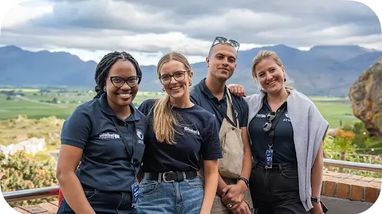 Four young adults wearing matching navy blue "Global Changemakers" shirts and event lanyards stand close together and smile for a photo. They are outdoors on a brick overlook with a vast green valley and layered mountain range under a cloudy sky behind them.