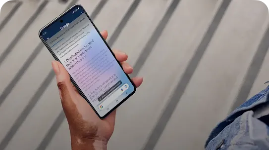A close-up of a person's hand with vitiligo holding a smartphone. The screen displays an article on Google titled '5 Mobile Management Trends,' and a prompt at the bottom reads 'Circle or tap anywhere to search.'