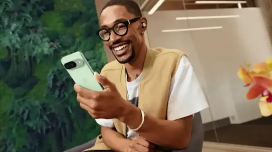 Man is sitting down in a chair and smiling while looking at his Pixel 9 phone.