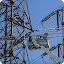 Two utility workers in a white cherry picker bucket performing maintenance on a large metal high-voltage power line tower against a clear blue sky.
