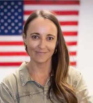 A woman with long brown hair smiling in front of a blurred American flag background, wearing a casual olive-green button-down shirt.
