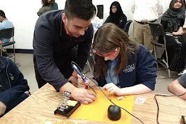 Thimble.io founder Oscar Pedroso assists a student as she practices soldering an electronic component onto a circuit board during a STEM workshop.