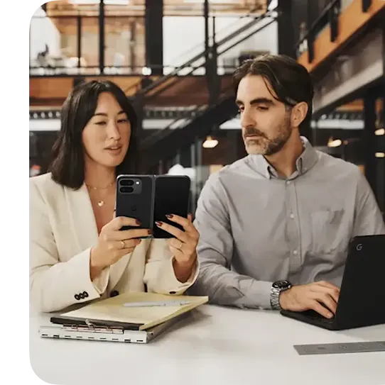 Two people seating at a desk in a modern office, looking at the phone together while working on laptops and notebooks.
