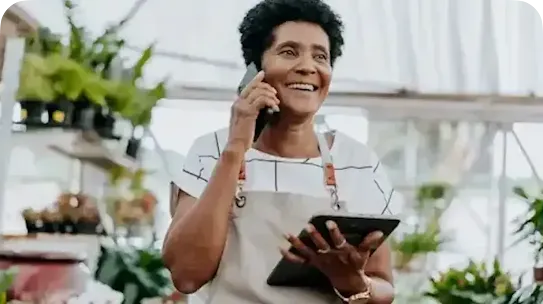A smiling woman with short curly hair, wearing a beige apron over a patterned shirt, talks on a smartphone while holding a tablet in a greenhouse filled with plants.