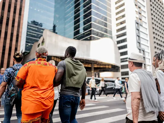 A diverse group of people walking across a busy city street with tall glass skyscrapers in the background.