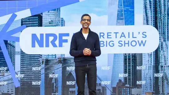 Sundar Pichai, CEO of Google and Alphabet, standing on a red stage during a keynote at "NRF: Retail's Big Show." Behind him is a large screen displaying the NRF logo and a New York City skyline. He is wearing a dark navy zip-up sweater and black trousers, presenting on the AI platform shift in the retail industry.