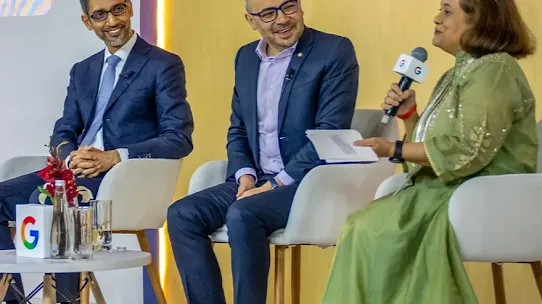 A panel discussion at the AI Impact Summit 2026. Sundar Pichai, CEO of Google and Alphabet, sits on the left in a dark suit, smiling as he listens to a woman in a green dress speaking into a Google-branded microphone. Another man in a navy blazer and glasses sits between them on a modern white stage against a warm yellow background.