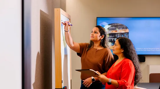 Two women are collaborating in a modern office or educational setting. One woman, wearing a brown t-shirt and dark trousers, stands at a framed whiteboard and uses a purple marker to draw a diagram. The second woman, dressed in a bright red blouse, stands beside her, holding a tablet and observing the board with a focused expression. In the background, a large digital screen displays a slide related to the Nobel Prize in Physics. The room is well-lit, featuring neutral-toned walls and professional furniture that suggests a brainstorm or academic discussion.