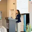 A woman standing at a podium giving a professional presentation about Gemini, Google's AI assistant.