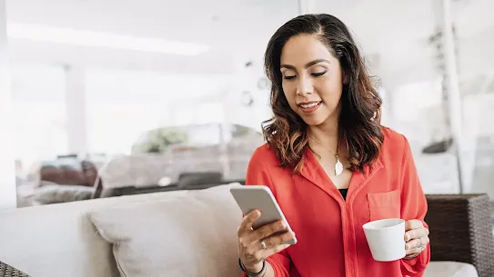 A woman in a n orange shirt sitting on a couch, holding a cup of coffee and looking at her smartphone.