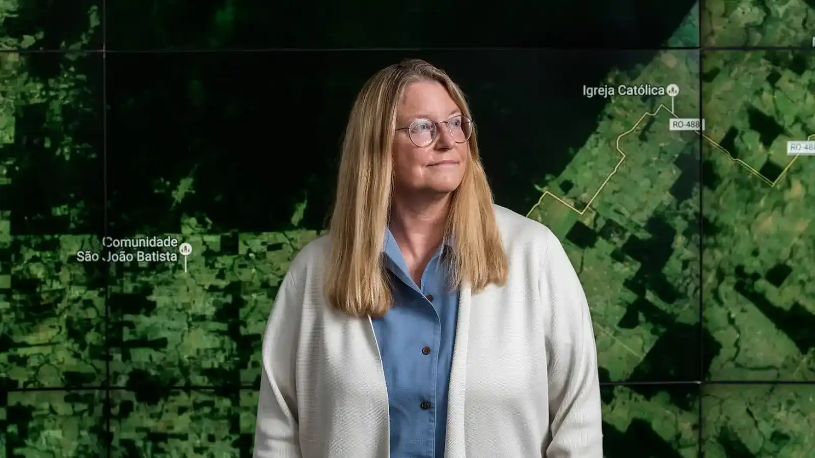 A woman with glasses wearing a blue shirt and white cardigan standing in front of a map, looking off to the left