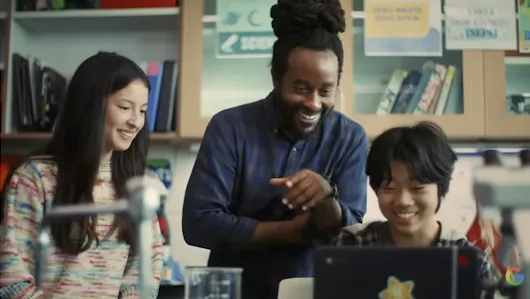 A smiling male teacher with dreadlocks points toward a laptop screen while working with two young students, a girl and a boy, in a science classroom. The background shows laboratory shelves with binders and educational posters, and a small Google "G" logo is visible in the bottom right corner.