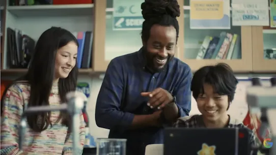 A smiling male teacher with dreadlocks points toward a laptop screen while working with two young students, a girl and a boy, in a science classroom. The background shows laboratory shelves with binders and educational posters, and a small Google "G" logo is visible in the bottom right corner.