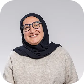 A smiling woman with glasses wearing a black hijab and a beige knit sweater, posed against a neutral grey background for a professional headshot.