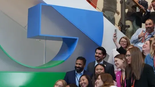 A group of smiling educators, the 2026 National Teachers of the Year, posing for a large group selfie on a staircase inside a Google office. They are gathered in front of a giant, multi-story Google "G" logo, with several people holding up phones to capture the moment.