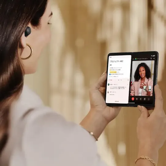A woman, in an office setting holding a mobile device, is on a video call with another female colleague.