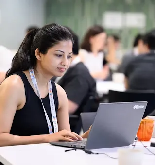 A woman wearing a "Google for Startups" lanyard working on a laptop at a shared desk in a busy workspace.