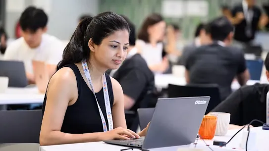 A woman wearing a "Google for Startups" lanyard working on a laptop at a shared desk in a busy workspace.