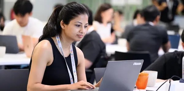 A woman wearing a "Google for Startups" lanyard working on a laptop at a shared desk in a busy workspace.