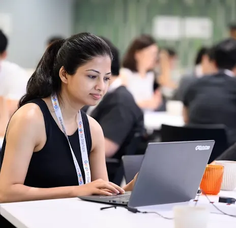 A woman wearing a "Google for Startups" lanyard working on a laptop at a shared desk in a busy workspace.