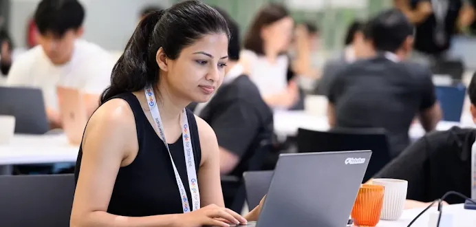 A woman wearing a "Google for Startups" lanyard working on a laptop at a shared desk in a busy workspace.