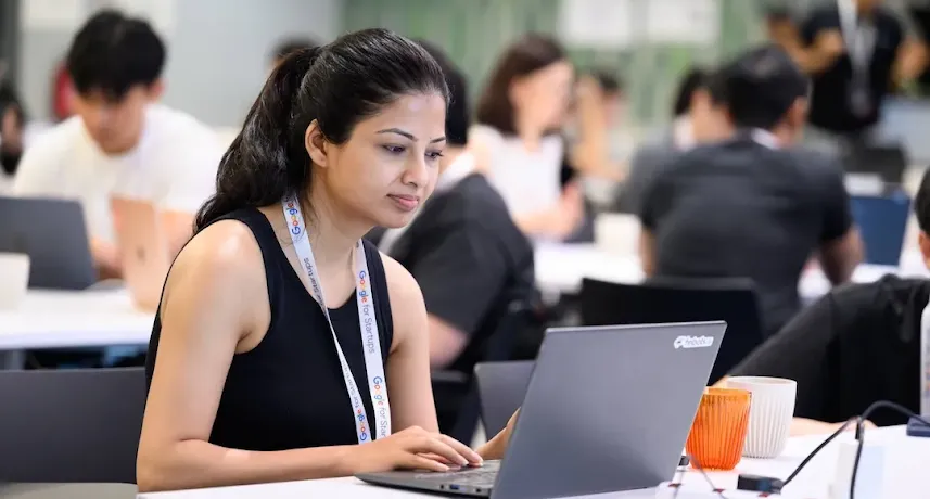 A woman wearing a "Google for Startups" lanyard working on a laptop at a shared desk in a busy workspace.